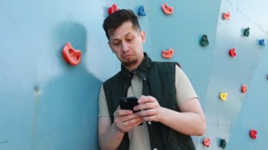 Bir adam tırmanma duvarının önünde cep telefonu tutarken görülüyor. Cihazla meşgul gibi görünüyor, muhtemelen mesajları kontrol ediyor ya da fotoğraf çekiyor..