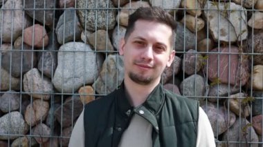 Gündelik siyah-beyaz bir kıyafet giymiş memnun bir genç adam, çeşitli taşlarla dolu tel örgülü bir çitin yer aldığı kendine özgü bir arka planın önünde hoş bir gülümsemeyle duruyor.
