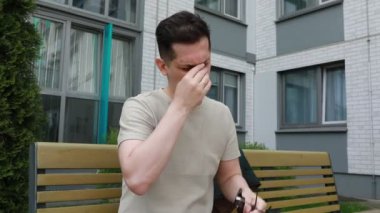 Bir adam bir bankta oturuyor, elinde bir makas tutuyor. Makasa odaklanmış görünüyor, muhtemelen kullanmaya hazırlanıyor. Arka plan, ağaçlı ve ağaçlı tipik bir açık hava ortamını gösteriyor.