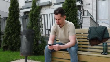 Bir adam bankta oturuyor, cep telefonunun ekranını dinlerken dikkati dağınık bir şekilde ona bakıyor. Cihazda her ne yapıyorsa ya da okuyorsa kendini kaptırmış görünüyor..