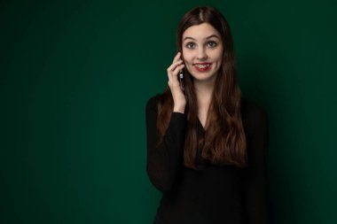 Bir kadın aktif olarak cep telefonuyla bir konuşma yapıyor, cihazı odaklanmış bir ifadeyle kulağına dayıyor. Halka açık bir yerde duruyor gibi görünüyor, muhtemelen sokakta ya da sokakta.