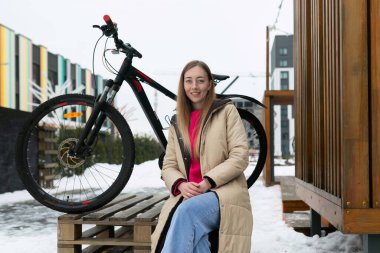 Tahta bir bankta oturan bir kadın, bir bisikletin yanına yerleştirilir. Kadın rahatlamış görünüyor ve potansiyel olarak ara veriyor. Bisiklet bankın karşısında duruyor, bu da onun tarzı olduğunu gösteriyor.