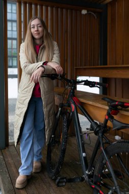 Dışarıdaki ahşap platformda bisikletli bir kadın. Kadın günlük kıyafetler giyiyor ve görünüşe göre bir mola veriyor. Bisiklet onun yanına park edilmiş ve ayarı kırsal..