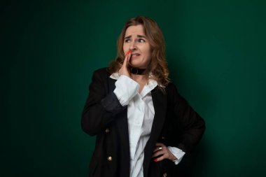 Bir kadın düz yeşil bir duvarın önünde duruyor, dümdüz ileriye bakıyor. Sakin ve sakin görünüyor, günlük giysiler giyiyor..