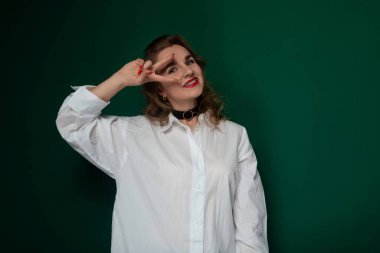 Beyaz gömlek giyen bir kadın fotoğraf için poz veriyor. Doğrudan kameraya bakarken kendinden emin ve rahat görünüyor. Duruşu zarif ve dengeli. Görsel olarak...