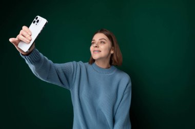 Mavi kazak giymiş bir kadın elinde cep telefonu tutarken ayakta duruyor. Ekrana odaklanmış gibi görünüyor, muhtemelen mesajlaşıyor ya da geziniyor. Arka plan basit,