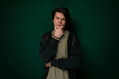 Katı yeşil bir duvarın önünde dimdik duran bir adam. Sıradan bir kıyafet giyiyor ve önüne bakıyor..