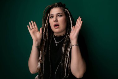 Titrek yeşil bir duvarın önünde kendinden emin bir şekilde duran rastalı bir kadın. Renkli saçları arka planda göze çarpıyor, çarpıcı bir görsel karşıtlık oluşturuyor..