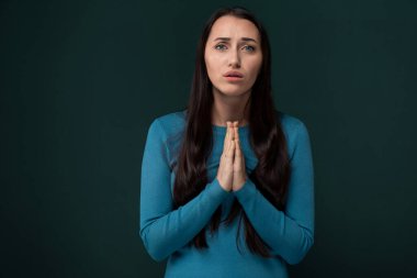 Mavi gömlek giyen bir kadın diz çökmüş dua ederken görülüyor. Elleri önünde birbirine kenetlenmiş ve gözleri konsantre bir şekilde kapalı..