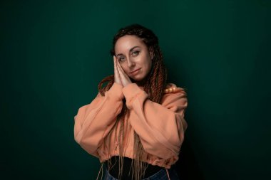 Rasta saçlı bir kadın parlak yeşil bir duvarın önünde duruyor. Benzersiz saç stiliyle kendine güvenen ve stil sahibi görünüyor. Yeşil arka planı da bireyselliğini vurguluyor..