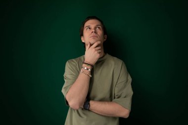 Katı yeşil bir duvarın önünde duran bir adam. Doğrudan kameraya bakıyor gibi görünüyor, kolları yanda. Yeşil duvar basit bir arkaplan görevi görüyor.