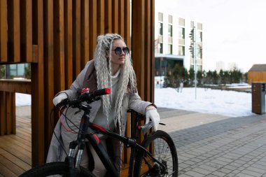 Bir kadın kaldırımda bisikletinin yanında duruyor. Günlük giysiler giyiyor ve ata binmeye ara vermiş gibi görünüyor. Kaldırımda arka planda binalar ve ağaçlar var..