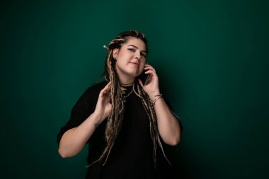 Rasta saçlı bir kadın aktif olarak bir telefon görüşmesi yapıyor. Kulağına bir cep telefonu dayamış ve yüzünde odaklanmış bir ifadeyle konuşuyor..