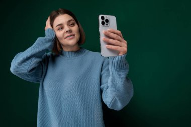 Bir kadın önünde bir telefon tutuyor, selfie çekerken kameraya gülümsüyor. Açık havada gibi görünüyor, yüzünde güneş ışığı var..