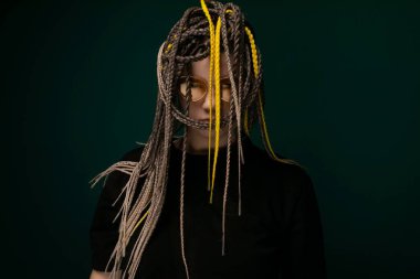 Rasta saçlı bir kadın, saf yeşil bir arka planın önünde duruyor. Kendine güveni tam ve doğrudan kameraya bakıyor..