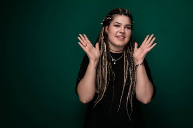 Rasta bukleli bir kadın sağlam yeşil bir zemine karşı duruyor. Kendinden emin görünüyor ve doğrudan kameraya bakıyor..