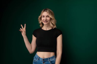 Bir kadın canlı yeşil bir duvarın önünde dikiliyor, doğrudan kameraya bakıyor. Kendinden emin ve sakin görünüyor..