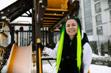 Canlı yeşil saçlı bir kadın renkli bir oyun alanının önünde kendine güvenerek kameraya doğru bakıyor. Etrafında salıncaklar, slaytlar ve diğer çocuk parkı ekipmanları var..