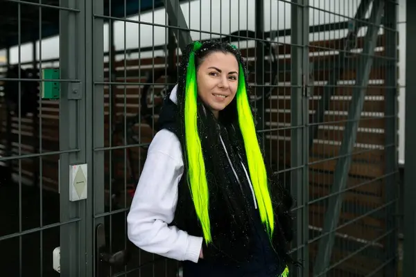 stock image A woman with vibrant green hair stands behind a metal fence, looking out into the distance with a thoughtful expression. The fence separates her from the open space beyond.