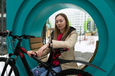 Mavi bir bisikletin yanındaki tahta bankta oturan kırmızı ceketli bir kadın. Arka planda ağaçlar ve binalar olan bir şehir parkı..