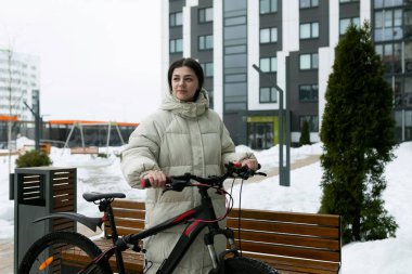 Karla kaplı bir arazide bir kadın bisikletin yanında duruyor. O, kalın giyinmiş, karlı manzaraya bakıyor. Bisiklet karlı zemine park edilmiş, etrafında kar taneleri yavaşça düşüyor..