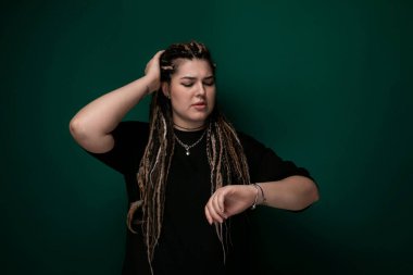 Titrek yeşil bir duvarın önünde kendinden emin bir şekilde duran rastalı bir kadın. Şık ve modaya uygun görünüyor. Kalın arka plana karşı eşsiz saç stilini sergiliyor..