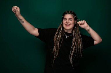 Rasta saçlı bir kadın canlı yeşil bir arka planın önünde duruyor. O, güvenle kameraya bakıyor. Yeşil arka plan onun koyu saçlarıyla çelişiyor, onu öne çıkarıyor..