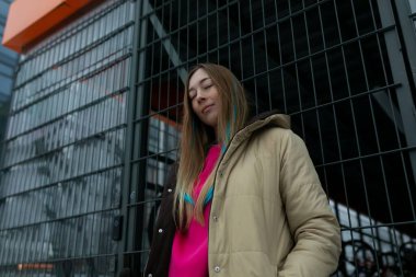 Modern, yüksek bir binanın önünde dikilip gökdelene doğru bakan sıradan giyinmiş bir kadın. Bina cam ve çelikten yapılmıştır. Çevredeki şehri yansıtır..