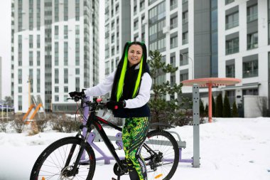 Karla kaplı bir arazide bir kadın bisikletin yanında duruyor. Kışlık giyinmiş ve bisikleti inceliyor gibi görünüyor..