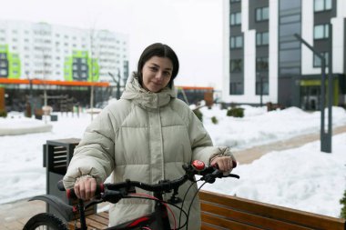 Karlı bir manzarada, bir kadın kalın giyinmiş, bir bisikletin yanında duruyor. Sahnede karla kaplı zemin ve bisiklete belirgin şekilde yer veriliyor.