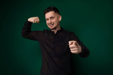 Siyah gömlek giyen bir adam ayakta duruyor ve fotoğraf için poz veriyor. Tarafsız bir ifadesi var ve doğrudan kameraya bakıyor..