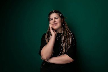 Rastalı bir kadın yeşil bir duvara yaslanıyor. Rahatlamış ve dalgın görünüyor, canlı arkaplana karşı duruyor..