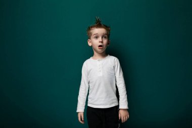 Genç bir çocuk canlı yeşil bir duvarın önünde duruyor. Yüzünde tarafsız bir ifadeyle dosdoğru karşıya bakıyor gibi görünüyor..