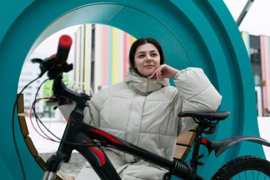 Bir kadın dışarıda bir sandalyede park edilmiş bir bisikletin yanında oturuyor. Uzaktan bir şeye bakarken rahatlamış görünüyor. Bisiklet çitlere yaslanıyor ve sahne basit ama huzurlu..