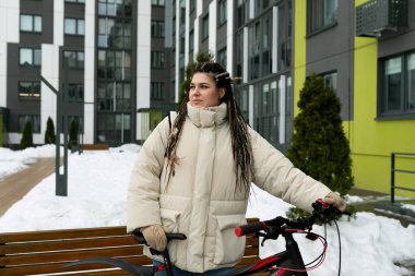 Rasta saçlı bir kadın şehir parkında bir bankın yanında duruyor. Uzağa bakarak dalgın dalgın görünüyor. Tezgah tahtadan yapılmış ve yıpranmış ama sağlam görünüyor..