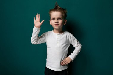 Genç bir çocuk canlı yeşil bir duvarın önünde duruyor, merakla çevresine bakıyor. Elleri yanı başında ve gözleriyle bölgeyi keşfediyor gibi görünüyor..