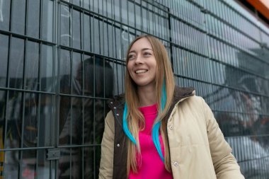 A woman is standing upright in front of a metal fence, looking straight ahead. She appears calm and composed, with no particular expression on her face. The metal fence is made of vertical bars and clipart