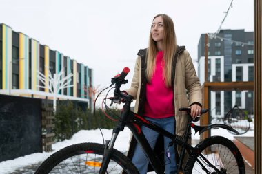 Bir kadın karda bir bisikletin yanında duruyor, gidona tutunuyor ve etrafına bakıyor. Kış kıyafetleri içinde, etrafında kar taneleri yağıyor..