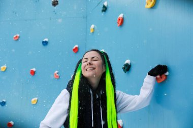 A woman with long hair is standing confidently in front of a challenging climbing wall, looking determined and focused. She appears ready to embark on a climbing adventure, surrounded by climbing clipart