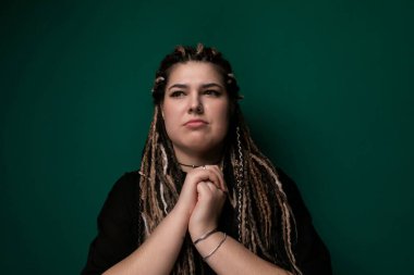 Rasta saçlı bir kadın canlı yeşil bir duvarın önünde kendinden emin bir şekilde duruyor. Saçları uzun, bükülmüş rastalarla kaplı ve doğrudan kameraya bakıyor..