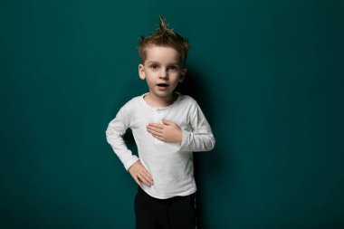 Genç bir çocuk canlı yeşil bir duvarın önünde duruyor, doğrudan kameraya bakıyor. Çocuk meraklı ve çevresiyle meşgul görünüyor, masumiyet ve merak hissi yaratıyor..