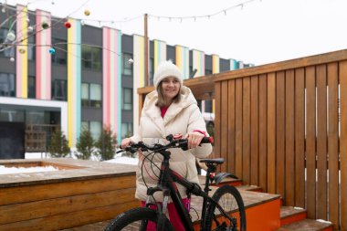 Bir kadın yüksek bir binanın önünde bir bisikletin yanında duruyor. Mola veriyor ya da talimatları kontrol ediyor gibi görünüyor. Bisiklet düzgün bir şekilde onun yanına park edilmiş ve o ileri bakıyor..