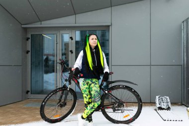 Gündelik kıyafetli bir kadın uzun bir şehir binasının arka planında bisikletin yanında duruyor. Yola çıkmadan önce duraklıyor ya da motoru güvenli bir şekilde kilitliyor gibi görünüyor..