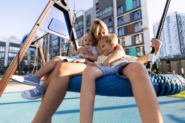 Bir anne, bir çocuk parkında birlikte sallanmaktan hoşlanan iki oğlunu nazikçe kucaklar. Parlak, güneşli bir gün, şehir parkında neşeli bir atmosfer yaratır, kahkaha ve bağ ile dolu. Şey...