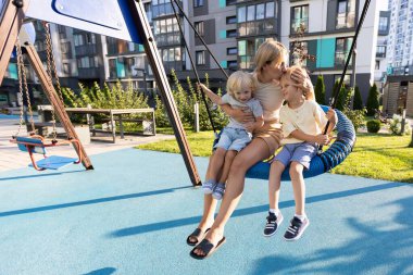 Bir anne, iki küçük oğluyla salıncakta oturup oyun parkında güneşli bir günün tadını çıkarır. Yeşillik ve modern binalarla çevrili olarak birlikte oynarken kahkaha ve gülücükleri paylaşıyorlar. Hayat dolu.