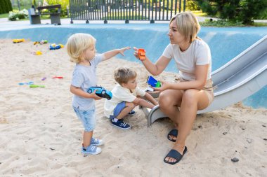 Bir anne, iki küçük oğluyla etkileşime girerken oyun parkında oturuyor. Oğullardan biri heyecan göstererek oyuncağa uzanırken, diğeri kaydırağın yanında oynuyor. Oyun alanı ağzına kadar dolu.
