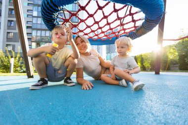 Neşeli bir anne ve iki oğlu oyun alanında birlikte eğleniyorlar. Renkli bir yapı altında eğlenceli bir şekilde etkileşime giriyorlar. Arka planda güneş batarken sıcak bir atmosfer yaratıyorlar.