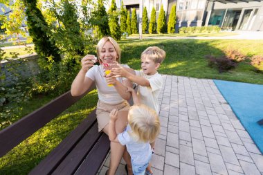 İki oğlu hevesle onunla ilgilenirken bir anne, canlı bir oyun parkında bir bankta oturuyor. Neşeli atmosfer, onların gülüşleri ve neşeli etkileşimleriyle güçlenir.