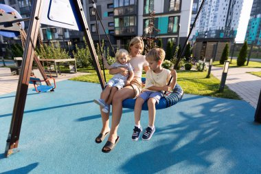 Bir anne, canlı bir oyun bahçesinde iki küçük oğluyla şakalaşarak sallanır. Modern binalar ve verimli yeşilliklerle çevrili, neşeli bir atmosfer yaratıyorlar. Kahkahalar havayı doldurur