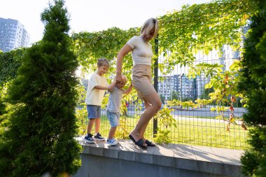 Bir anne, parlak ve neşeli bir oyun parkında alçak bir duvar boyunca neşeyle yürüyen iki küçük oğluna rehberlik eder. Çevreleyen yeşillik ve kentsel zemin eğlencelerine canlılık katıyor.